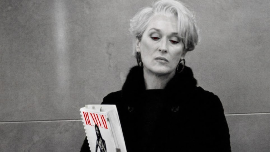 Una impactante interpretación visual de Miranda Priestly. En un mundo monocromático, su fría mirada brilla con un rojo penetrante, subrayando el poder absoluto que ejerce desde su trono en la revista RUNWAY. ¿Acaso ve un error de estilo o simplemente el futuro de la moda? La agenda que sostiene, con la palabra RUNWAY en rojo, es el mapa de su imperio.